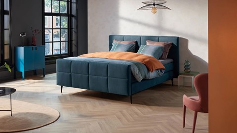 een blauw bed met oranje kussens en een zwarte plafondlamp