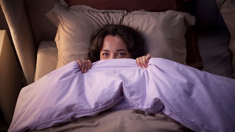 Een vrouw die in bed ligt en haar gezicht bedekt met een deken